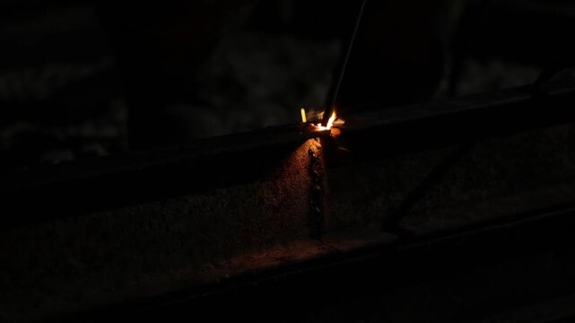 Close framing shows welder guiding electrode on rusted steel I beam as arc erupts, sparks fly, smoke curls, and molten droplets scatter under intense bright light.