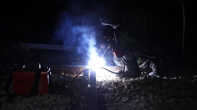 Helmeted worker kneels on gravel, operates torch with bright arc and smoke. Red welding units, cables, guard rail, and post mark scene during off peak repairs.