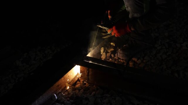 Worker in orange gloves kneels on ballast and performs arc welding on steel rails at night, bright white white arc emits smoke and sparks, cables and tools are visible