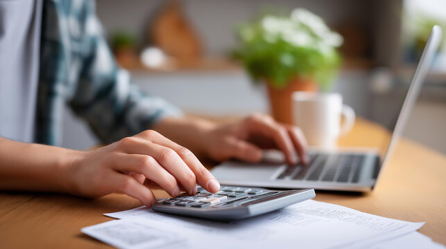 Hands holding a calculator beside an open laptop and paperwork on a kitchen table, defocused bills in the background, home finance, budgeting, financial planning, household economy, laptop work,