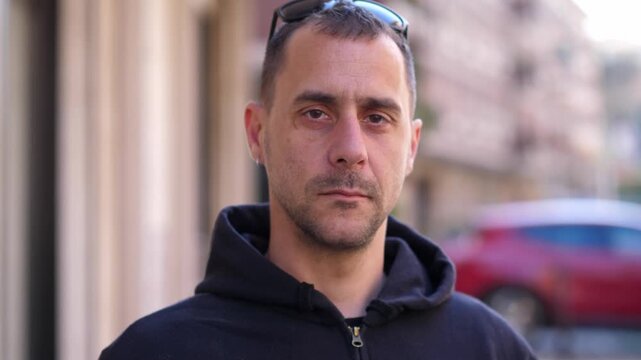 Young man in a black hoodie looks straight into the camera with a serious and impassive expression. The scene conveys intensity and emotion control.