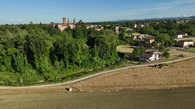 Harvested field with tractor forming hay bales near Castello di Castelnuovo Fogliani in Castelnuovo Fogliani, Emilia-Romagna, Italy, with rural farmland, trees and village buildings in Po Valley lands