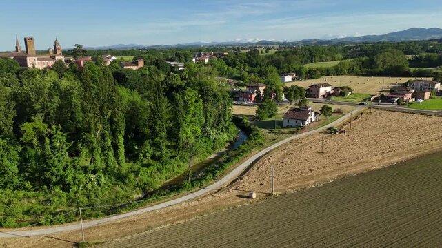 Aerial view of harvested field with tractor forming hay bales near Castello di Castelnuovo Fogliani in Emilia-Romagna, Italy, with rural landscape, drone orbiting