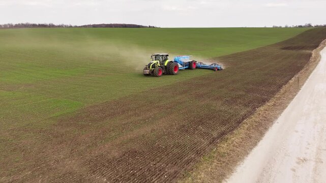 Green telehandler lifts a blue tank while a large plow is attached, set in a farm field with brown soil and green grass in the background