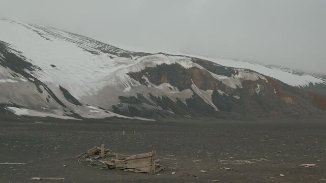Collapsed decayed wooden boat hull lies abandoned on dark volcanic ash plain at Whalers Bay, Deception Island, with dramatic snow-streaked volcanic slopes rising behind