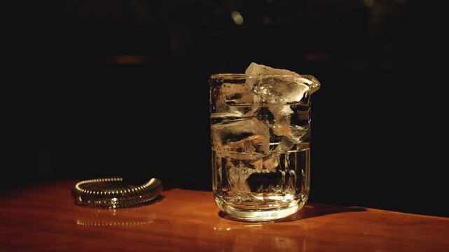 Clear ice cubes in mixing glass on wooden counter with dark atmospheric lighting for craft cocktail preparation.