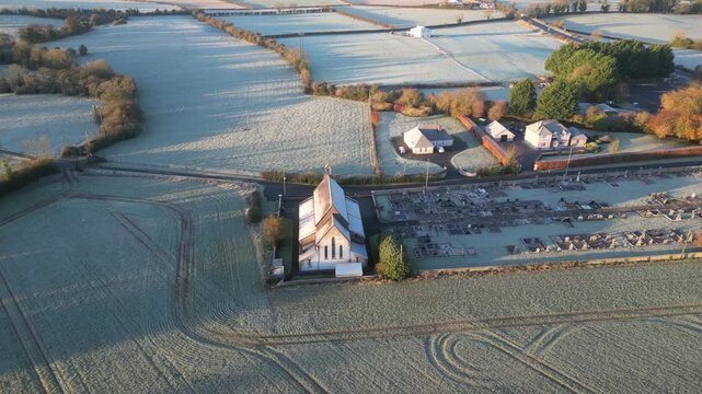 High-altitude drone video captures Saint Brendan's Church, graveyard, and frosty fields in County Kilkenny at sunrise. Aerial footage highlights the serene winter landscape.