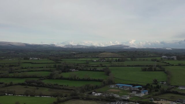 High-angle aerial view of Suir Valley in Portlaw, Ireland, showcasing lush green fields and distant snow-capped mountains under a cloudy sky, offering serene rural beauty.