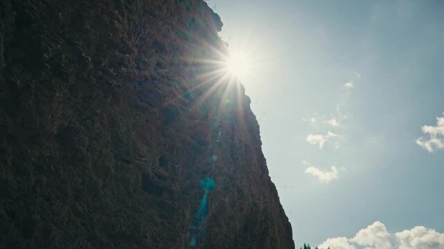 Bright sun star peaks from behind a cliff against a clear blue sky, creating a dramatic light effect and highlighting natural contrast in a serene outdoor scene.
