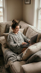 Obraz premium Young woman with hair bun sits on comfortable sofa with laptop. She is smiling while typing. Cozy home atmosphere with soft light. Ideal for remote work or lifestyle content.