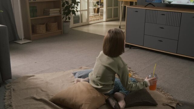 Full dolly-in shot of 8-year-old Caucasian girl running into living room, sitting on blanket and pillows on floor to watch video game stream on TV, while snacking on chips and drinking juice