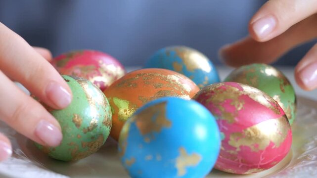 Hands gently pick up colorful, gold-embellished eggs. Each egg has unique, vibrant paint patterns. They rest on a white plate in close-up. Background softly blurred for focus on eggs and hands