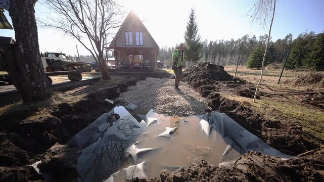 Low angle wide shot shows sun flare over roof, pond liner holding muddy water, long crushed stone path, worker in high visibility jacket, fence and conifer treeline.