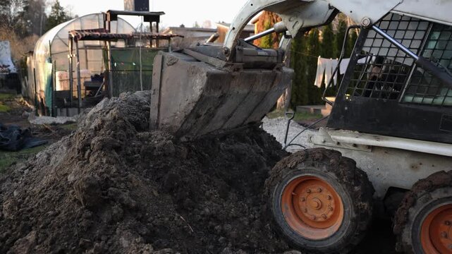 Skid steer loader digs in a residential yard, mud caked bucket scoops dark soil into a mound as tracks press into ground, with clothesline and wooden swing nearby.