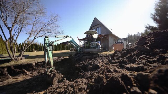 Compact excavator lifts soil near a triangular roof house as a dog moves. One operator works by hedgerow and leafless tree in daylight with wide lens and low view.