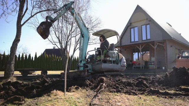 Small turquoise excavator lifts roots and piles soil at a steep gabled house. Tracked undercarriage shifts as operator runs controls. Midday light and low angle.