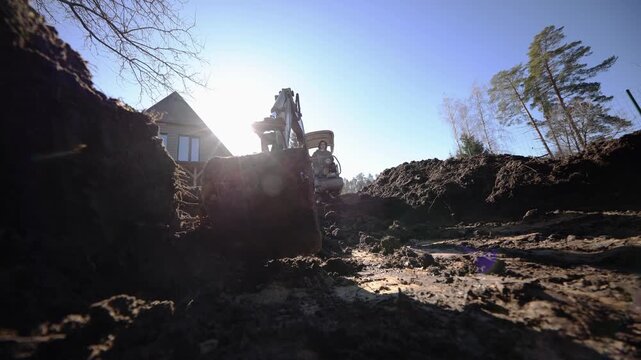 Low angle view shows mini excavator lifting bucket in a deep trench as tracks churn wet clumpy soil, throwing mud. Strong backlight rims the machine against sun.