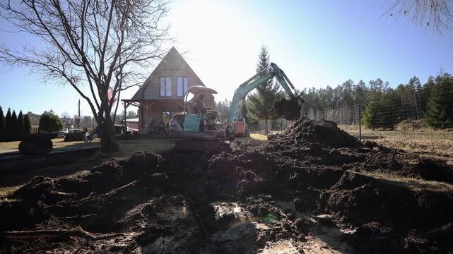 Ground level view shows tracked mini excavator scooping dark soil by a timber cabin with porch and leafless tree, operator in cab, muddy water, strong backlight.