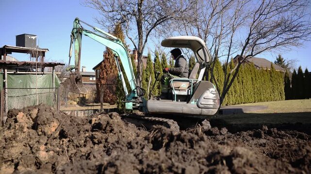 Small turquoise tracked excavator lifts and drops earth as flecks scatter, operator focuses on controls, wide shot, natural light, shallow depth, midmorning work.
