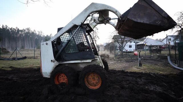 Skid steer loader lifts compacted soil as wheels sink in churned mud. Late winter light, low angle, cool muted tones, soft directional light, tight crop show motion.