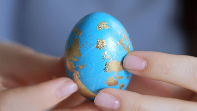 Close-up of hands holding a blue egg with gold patterns. The egg has natural speckles. It's a real bird egg. The egg is being gently turned. Background is softly blurred