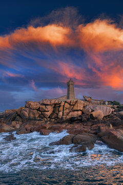 Aerial view of the Mean Ruz Lighthouse on the pink granite rocks of Ploumanac'h with crashing waves under a dramatic sunset sky in Perros-Guirec, Brittany, France.