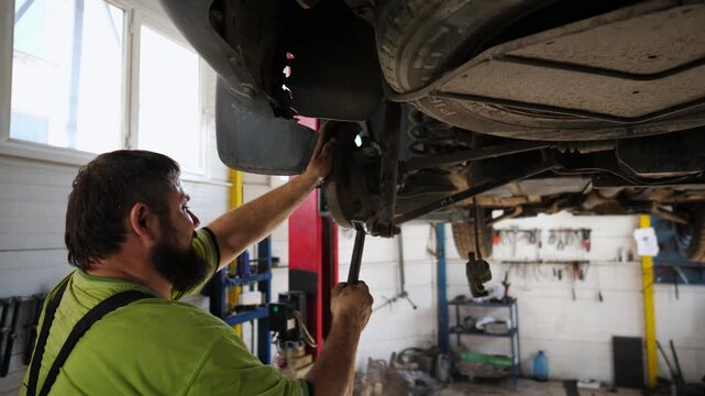 Professional bearded auto mechanic in uniform repairs detail of car using hammer at workshop. Adult male repairer works underneath a lifting vehicle at garage. Concept of automobile maintenance