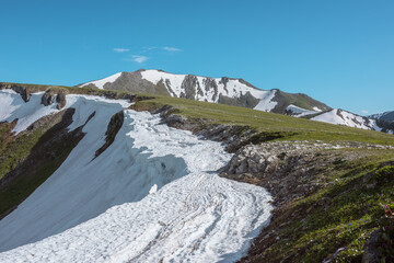Scenic alpine sunlit view along green grassy ridge with white snow cornice and rocky sheer crags in bright sun under blue sky. Awesome landscape with sharp rocks and snowy mountain range in sunny day. © Daniil
