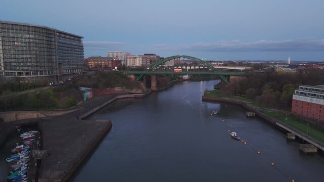 Aerial drone shot flying forward over the River Wear at dawn toward the Wearmouth Bridge and Monkwearmouth Railway Bridge, with the Sunderland city skyline in the background.
