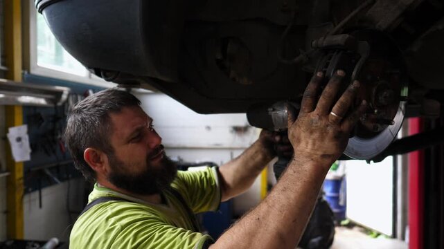 Male repairer tightening bolts with special electric tool from car detail at workshop. Professional auto mechanic works underneath lifting vehicle at garage. Adult repairman fixing transport indoor