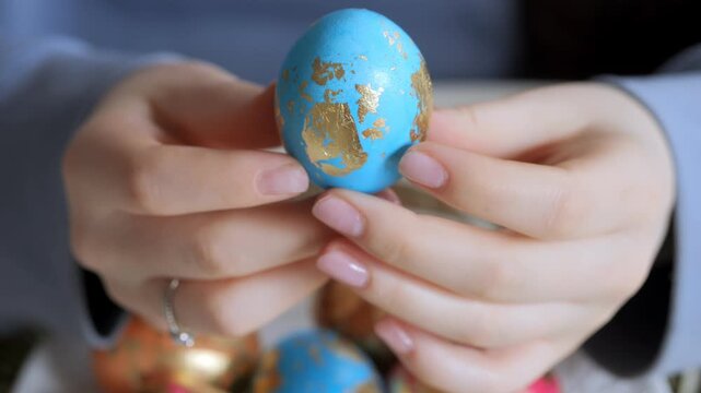 Hands hold blue egg with gold map design. Background shows more decorated eggs. Focus stays on egg's detailed pattern. Soft lighting highlights texture and color