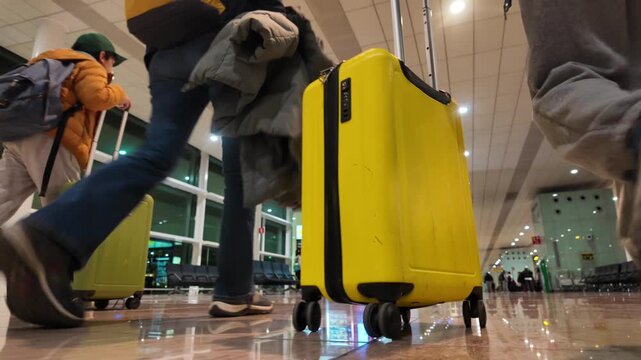 Low angle view of travelers legs walking with luggage and rolling a yellow suitcase through a modern airport terminal reflecting on the shiny floor, ready for departure