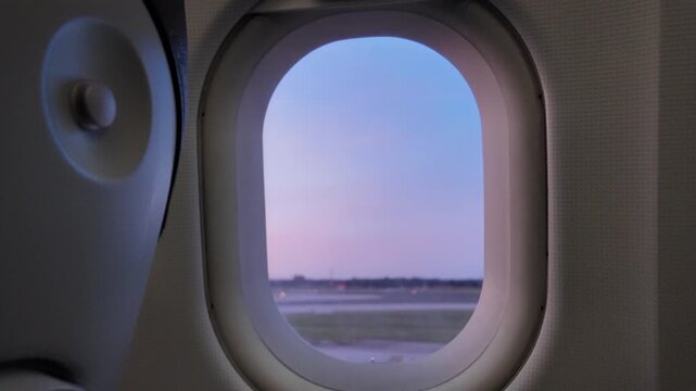 Airplane window view of the airport tarmac during a colorful sunset while preparing for takeoff, showing the wing and the runway lights before the flight