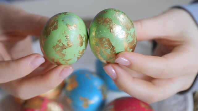 Hands hold two green and gold decorated Easter eggs. Background shows colorful eggs in soft focus. Close-up shot highlights intricate patterns on eggshells. Gentle lighting enhances festive