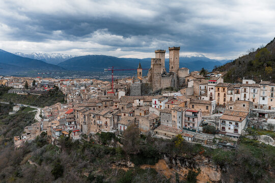 Aerial view of Caldora Castle and the medieval stone houses of the village under a dramatic cloudy sky with snow-capped mountains in the background Pacentro, Abruzzo, Italy.