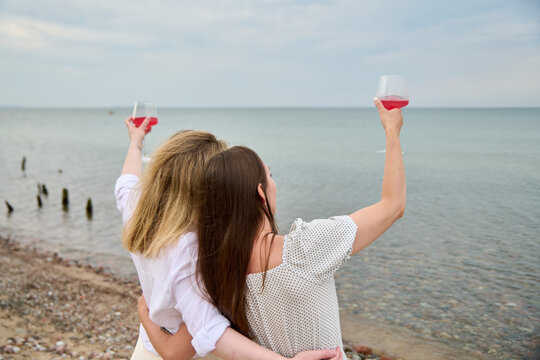 Two young women toast with pink wine at seaside picnic embracing friendship