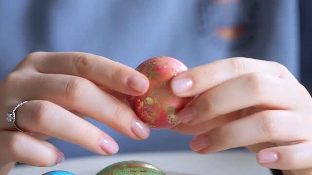 Hands hold decorated egg with red and gold pattern. Other colorful eggs visible nearby. Person wears ring and has manicured nails. Blurred background suggests indoor setting