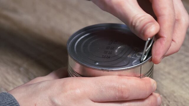 Man's hands opening a tin can of beans by pulling the ring pull tab on a kitchen table. Close-up of opening canned food.