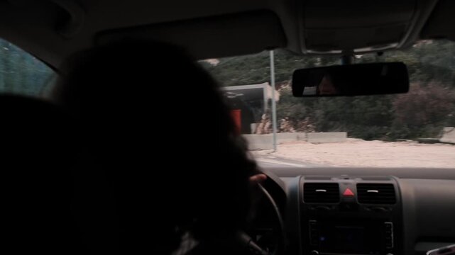 Woman driving car turning on road near checkpoint seen from interior. Driver silhouette behind steering wheel, dashboard view, vehicle moving on curved road in mountainous area.