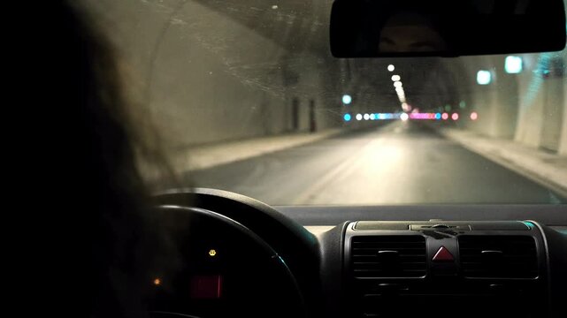Defocused view of road inside tunnel from car interior. Blurred lights, oncoming vehicle headlights and driver silhouette, dashboard perspective during night driving.