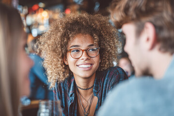 Fototapeta premium A young woman with curly hair and glasses smiles while talking with friends in a lively indoor setting.