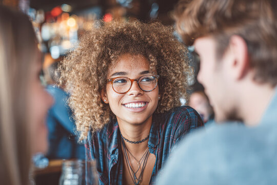 A young woman with curly hair and glasses smiles while talking with friends in a lively indoor setting.