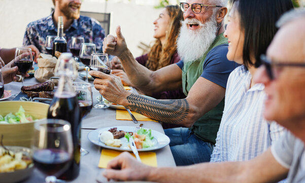 Happy family people having fun at barbecue lunch - Multiracial friends eating at bbq meal - Food, friendship, gathering and summer lifestyle concept - Focus on hipster man left hand