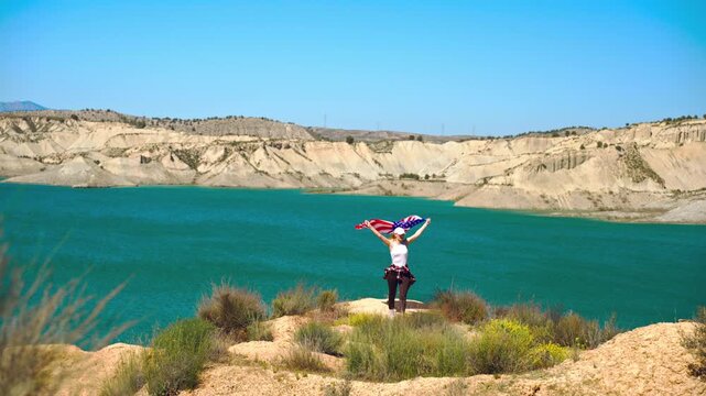 Young woman standing on mountain summit holding USA flag above peaceful lake, symbolizing inner strength, personal transformation, freedom and emotional breakthrough.