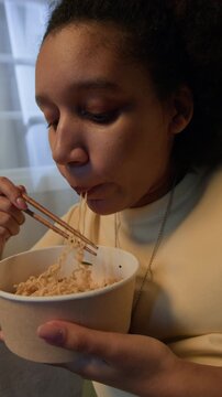 Vertical slowmo shot of young African American woman slurping noodles from takeaway bowl using chopsticks at home in evening