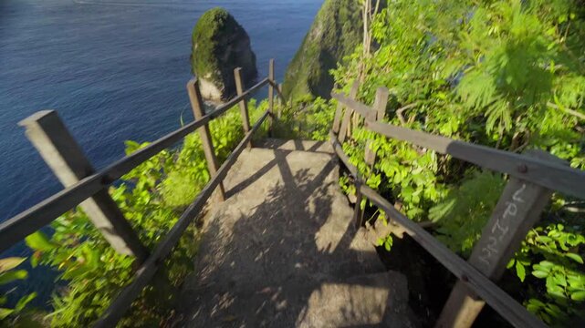 Man walking down steep trail and stairs to Kelingking Beach on Nusa Penida, Bali, surrounded by cliffs and ocean views.