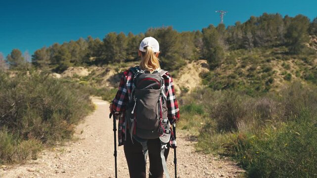 Active woman with backpack and trekking poles training endurance while walking on mountain dirt road in sunny weather. Concept of fitness, cardio training and outdoor sport activity
