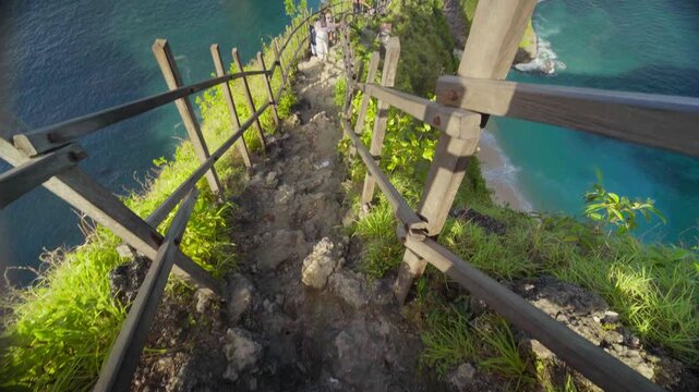 Man walking down steep trail and stairs to Kelingking Beach on Nusa Penida, Bali, surrounded by cliffs and ocean views.