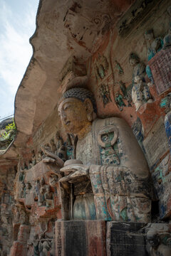 Ancient stone Buddha statue carved into cliff face. Weathered carvings surround serene seated figure. Vegetation peeks through cracks above carved surface. Faded colors remain on stone