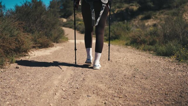 Woman hiking away from camera on a mountain dirt road with trekking poles, showing steady movement and outdoor endurance. Concept of journey, freedom, fitness and active lifestyle in nature.